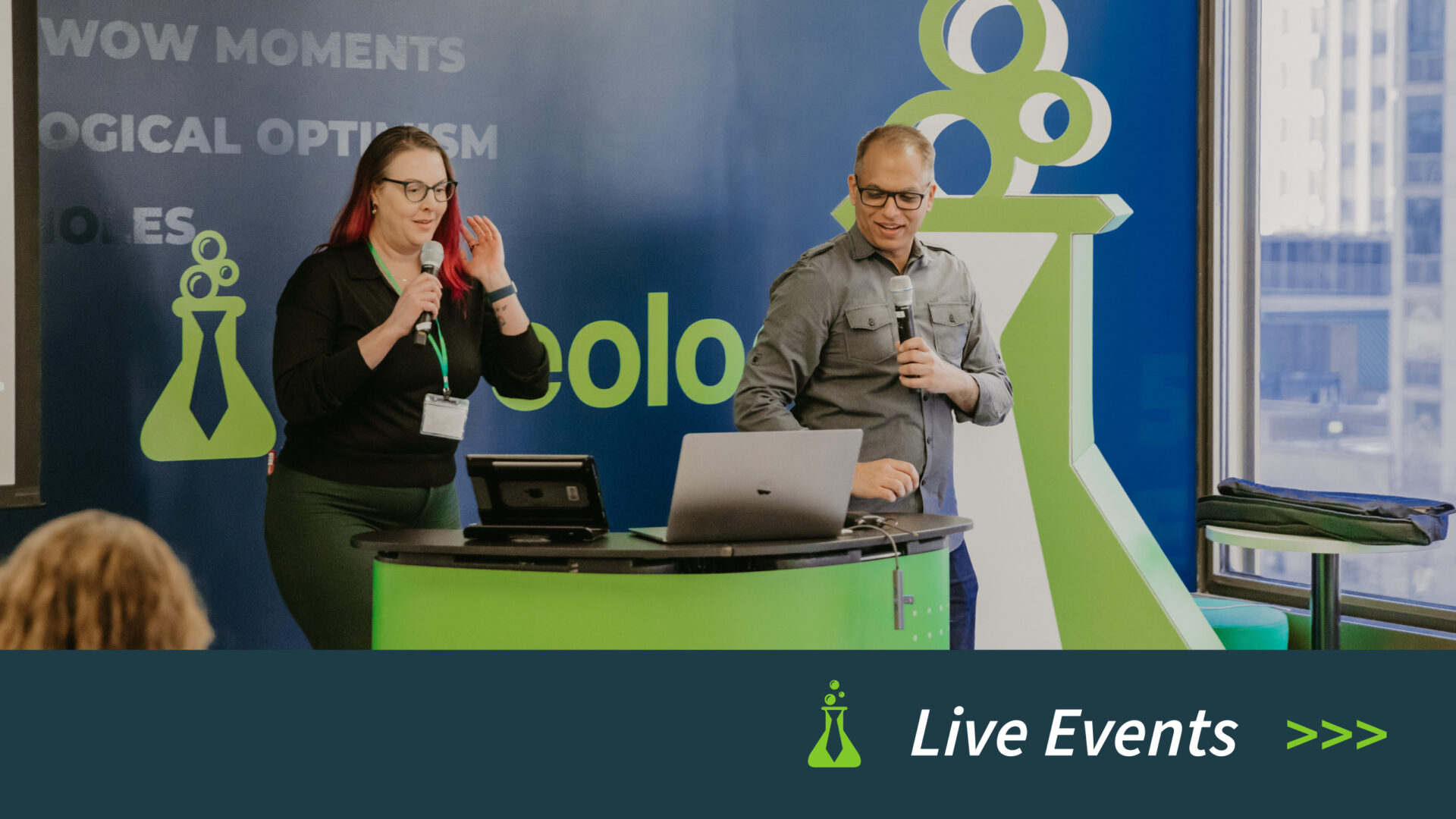 Two presenters with microphones at a green and black desk in front of a branded blue and green backdrop.
