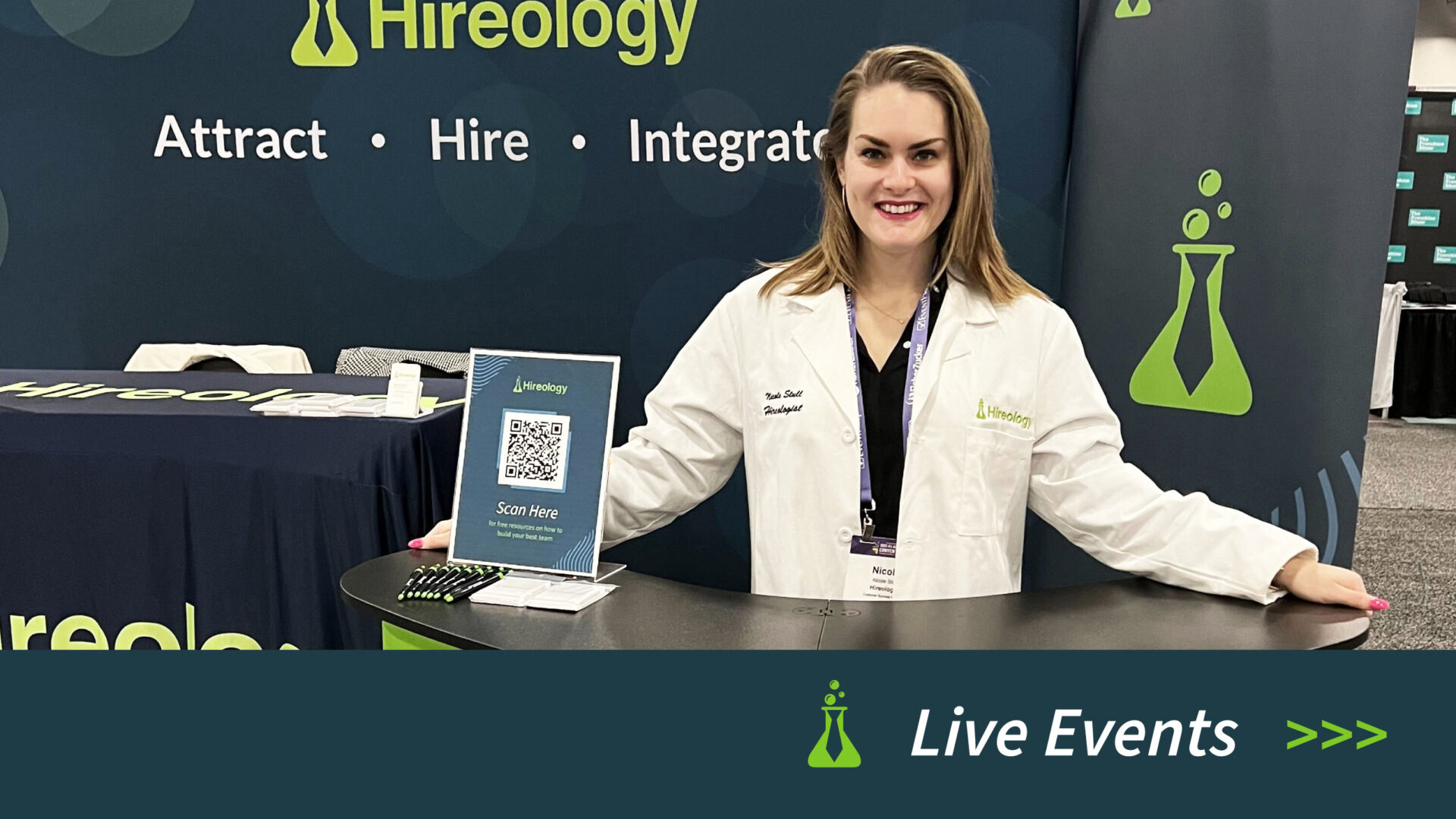 Smiling woman in white coat at Hireology booth with QR code sign for live events.