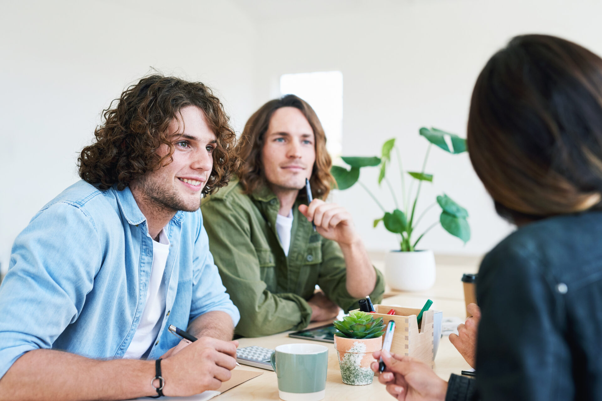Two men with curly hair listening attentively to a woman during a casual meeting with plants on the table.