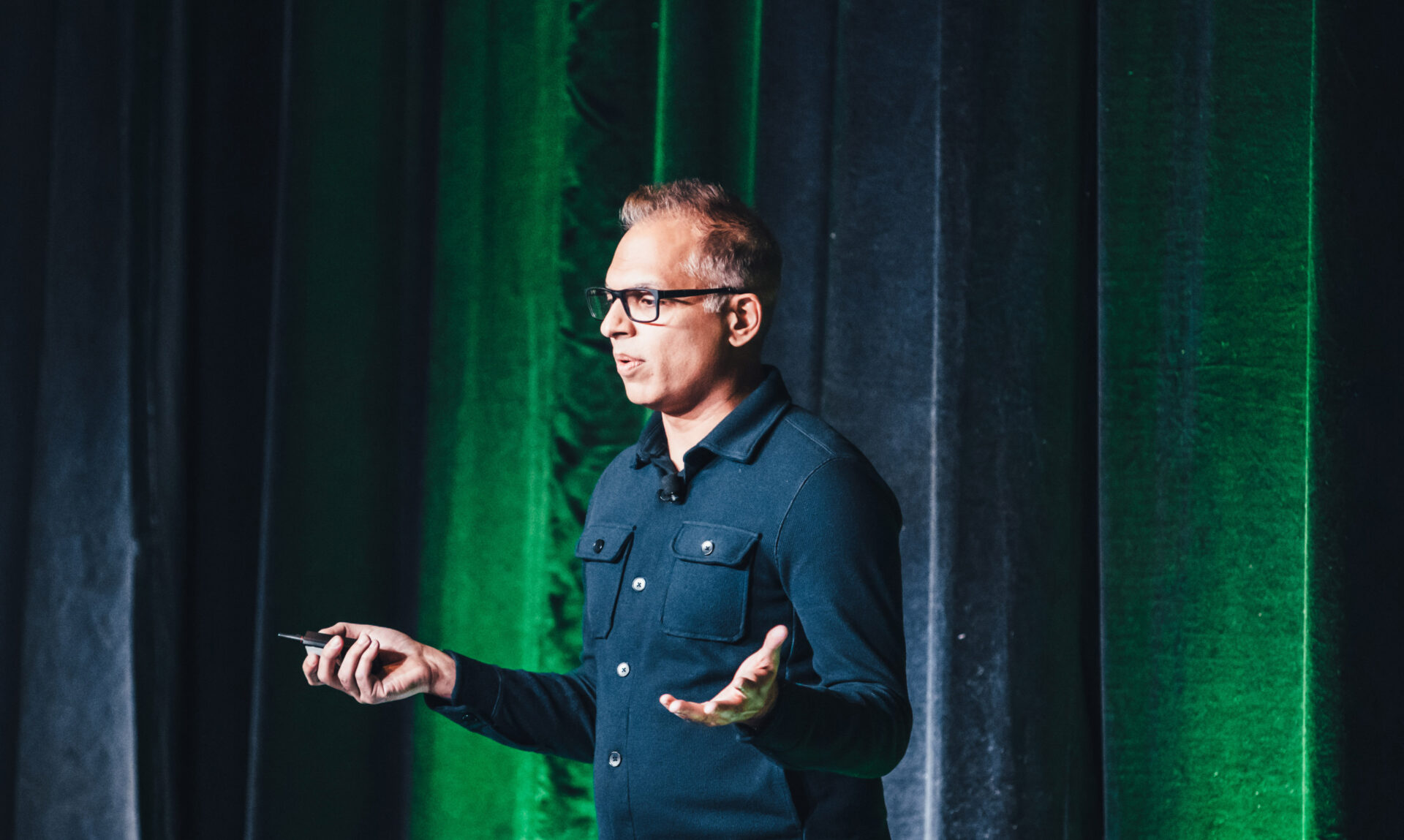 Man wearing glasses giving a presentation with green and dark curtains in the background.