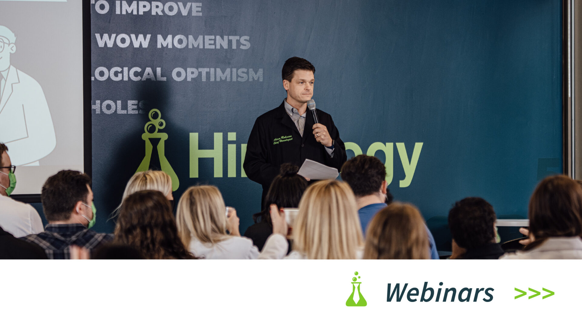 Man holding microphone and papers speaking to seated audience with webinar logo on blue wall behind him.