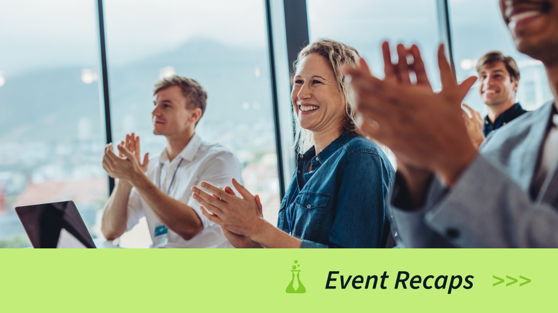 Smiling people sitting and clapping together at a conference or event in a bright room with large windows.