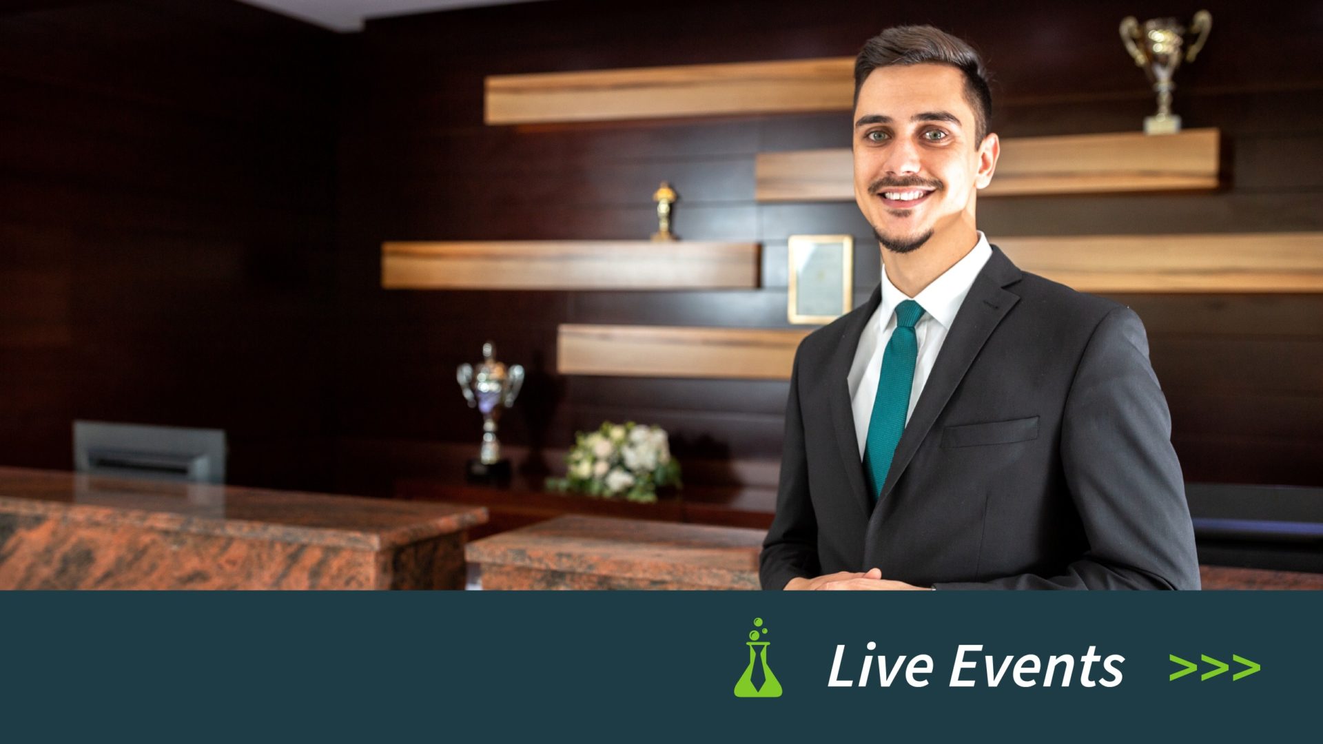 Smiling man in a suit and teal tie standing in a lobby with trophies on wooden shelves behind him.