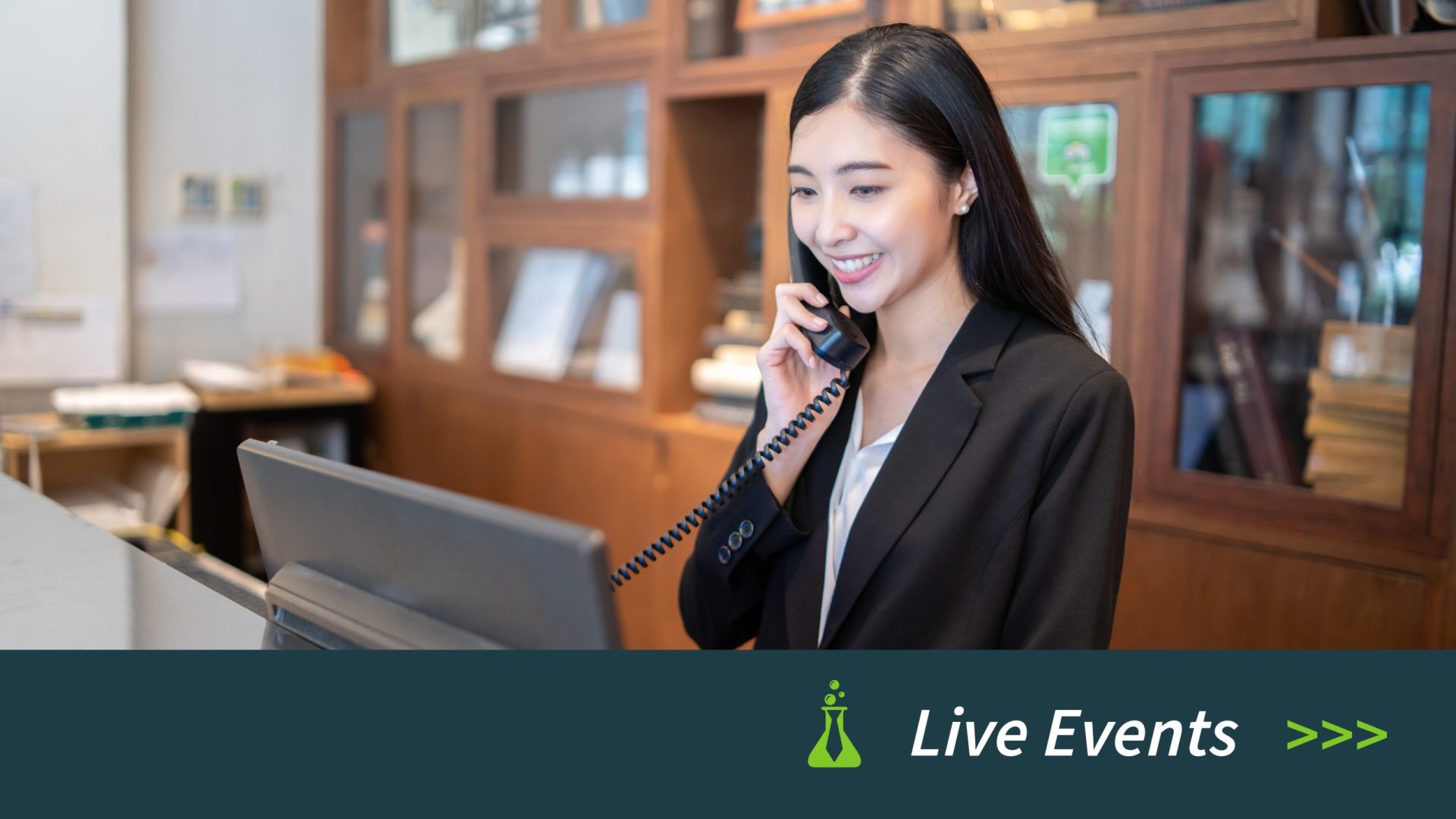 Smiling woman in black blazer using telephone at a counter with wooden shelves in the background.