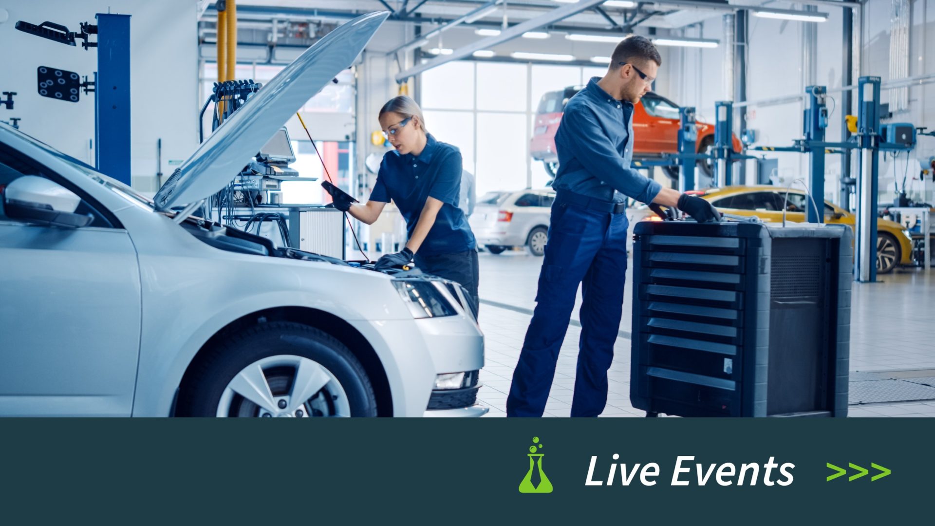 Two mechanics working on a silver car in a spacious, well-lit auto repair shop.