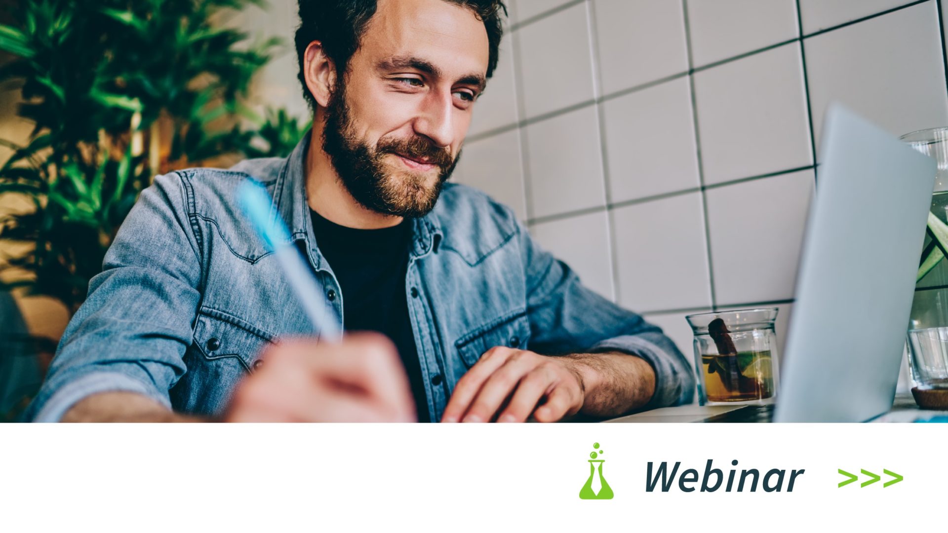 Man with beard writing in notebook while looking at laptop in a tiled room with plants.