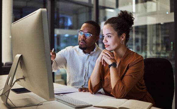 Two coworkers collaborate looking at a desktop computer in a modern office at night.