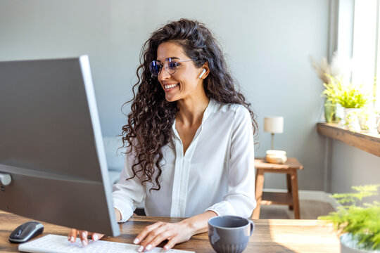 Smiling woman wearing glasses using desktop computer with wireless earbuds at wooden desk by window.