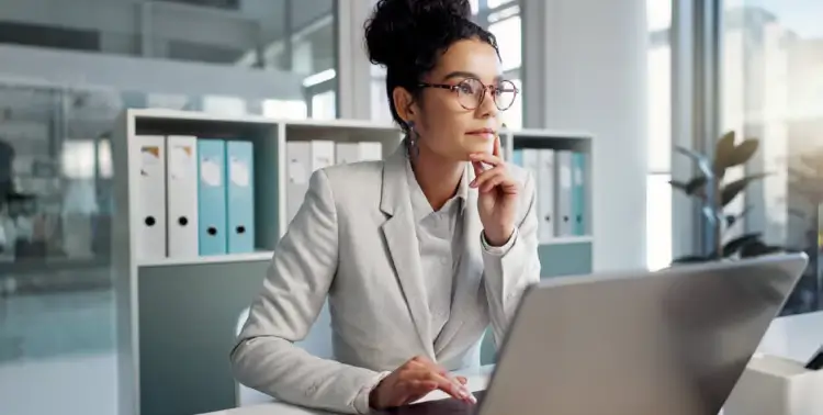 Woman in glasses and blazer thoughtfully working on a laptop in a modern office setting.