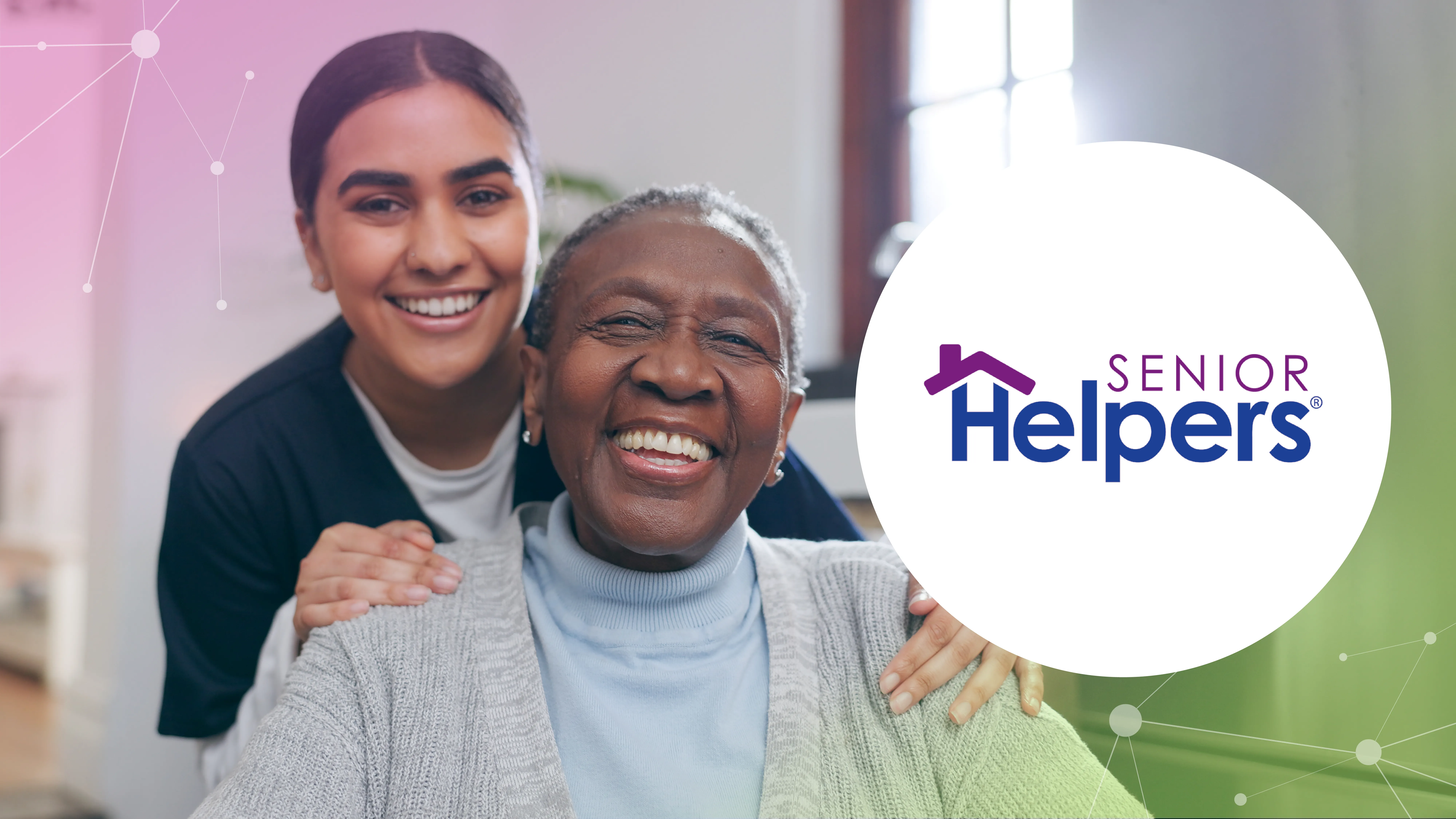 A smiling young caregiver stands behind a smiling elderly woman in a cozy indoor setting.