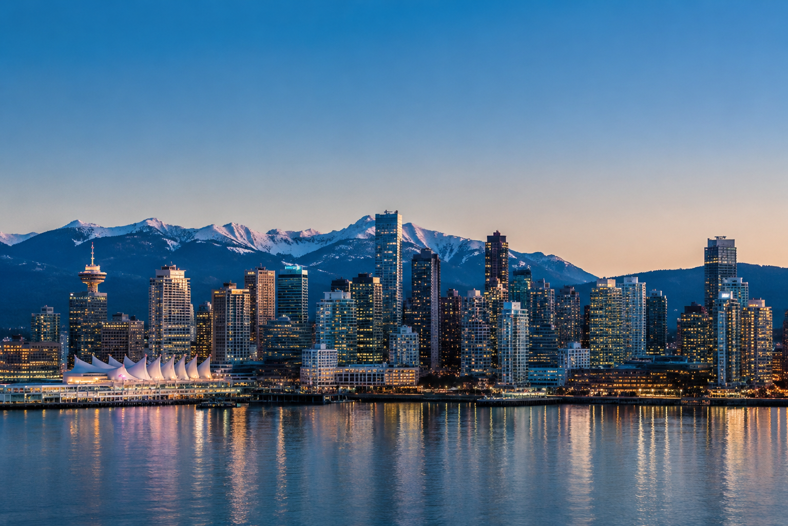 City skyline with tall buildings reflecting on water and snow-capped mountains under a clear sky at dusk - METRO VANCOUVER