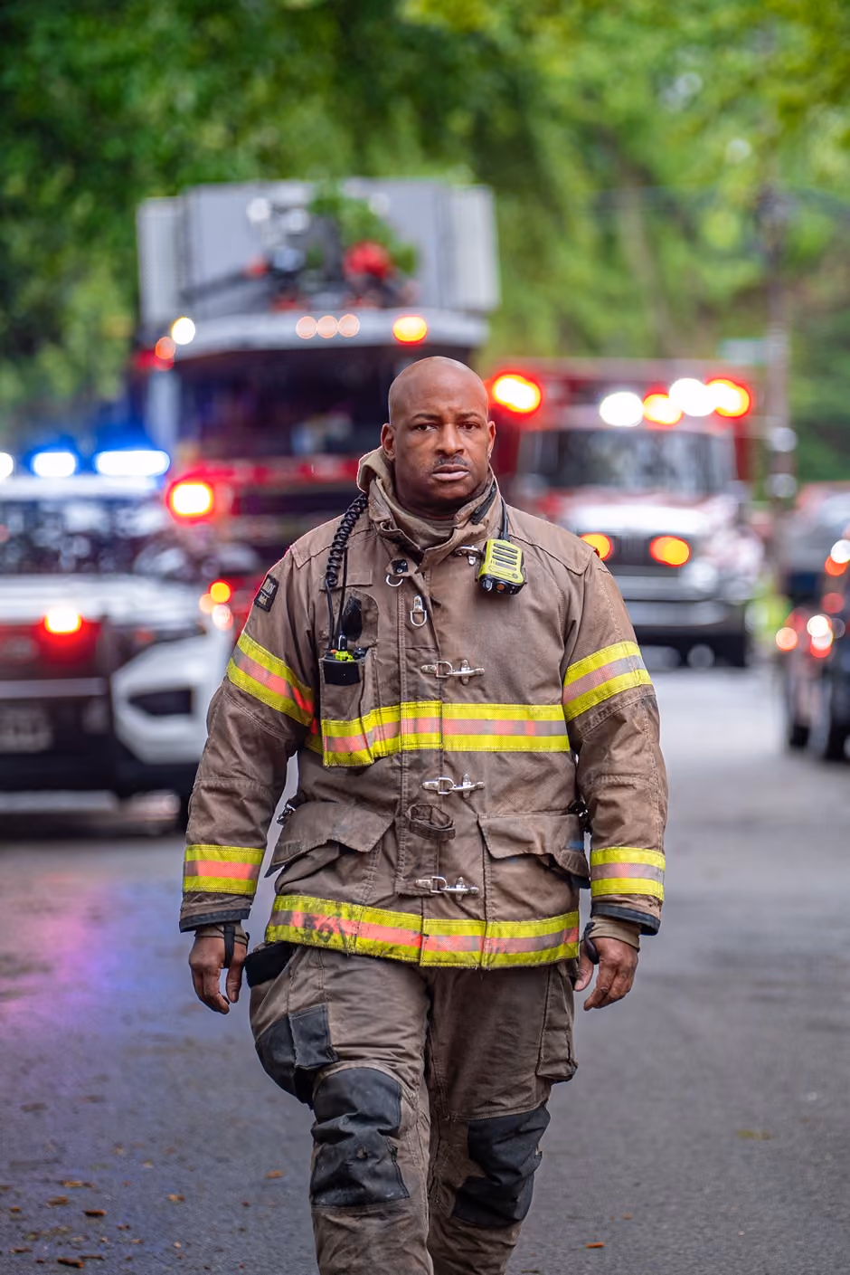 Firefighter in gear walking on a street with fire trucks and emergency lights in the background.