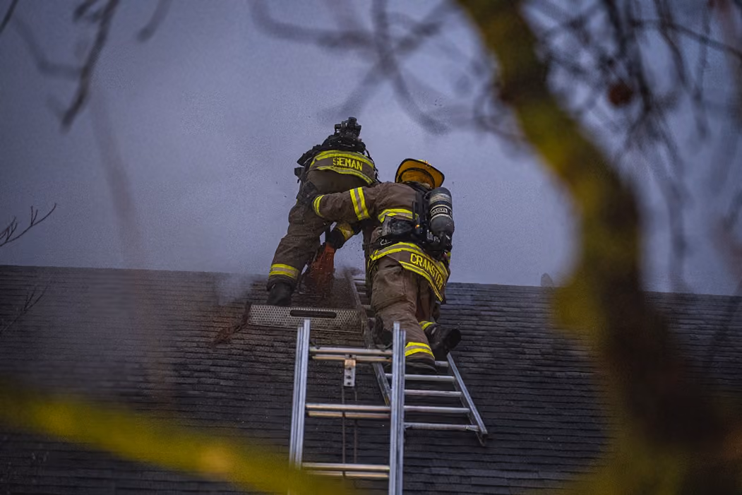 Two firefighters in gear climbing a ladder to a rooftop through smoke at dusk.