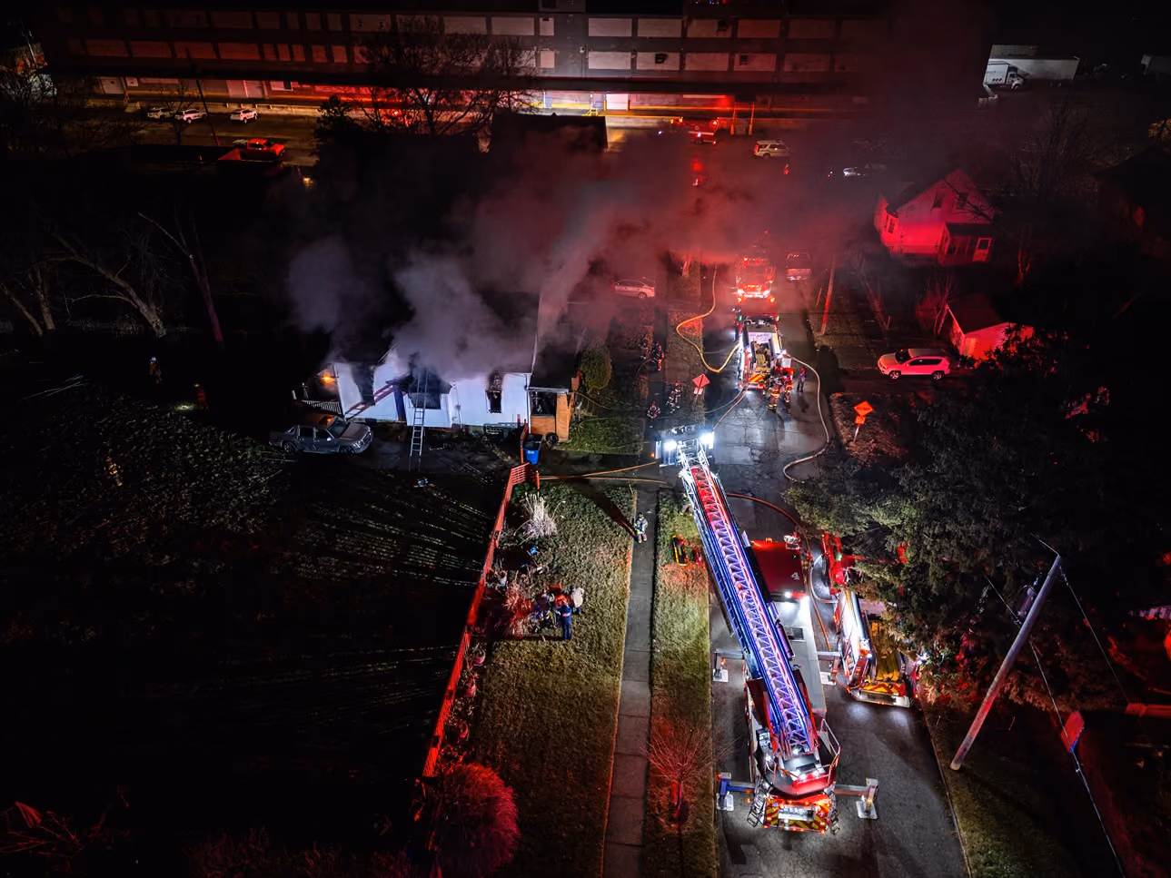 A nighttime aerial view of firefighters using a ladder truck to extinguish a house fire with smoke billowing out.