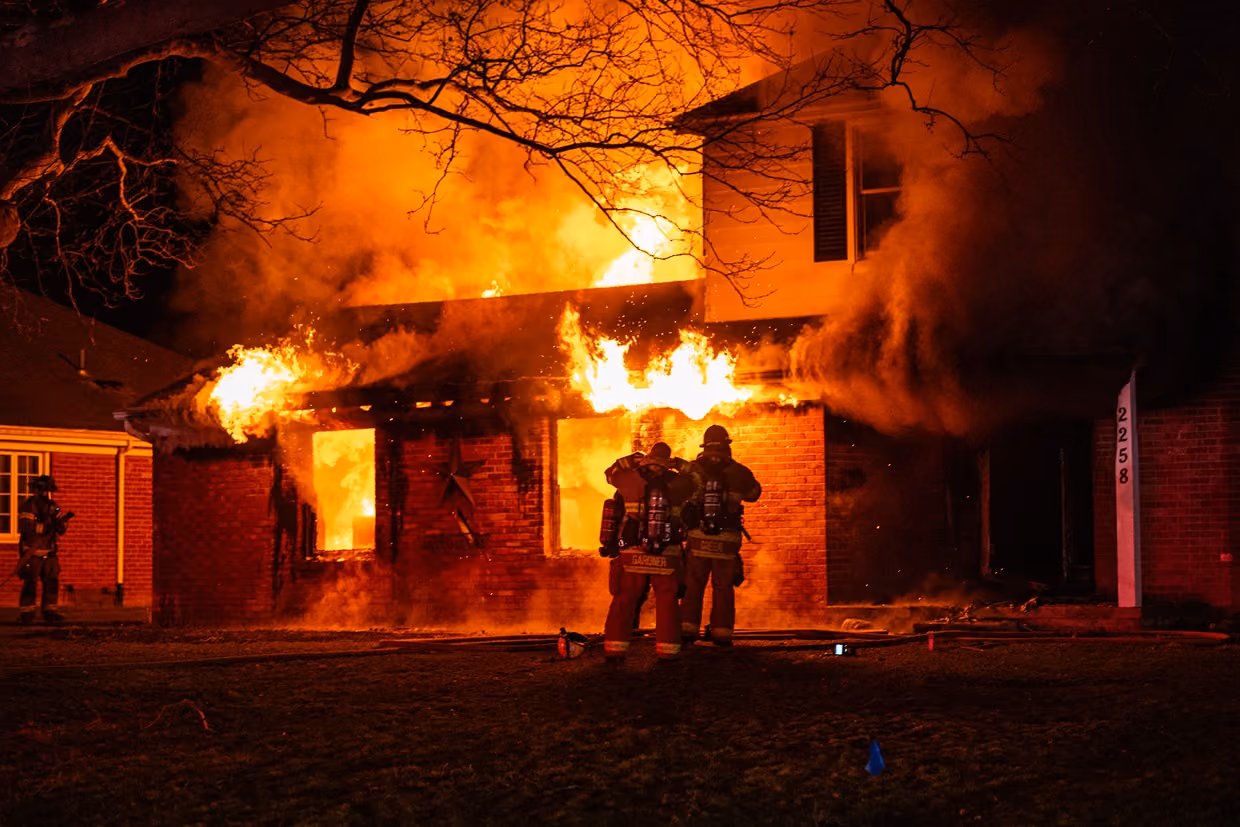 Two firefighters in gear standing in front of a house engulfed in flames at night.