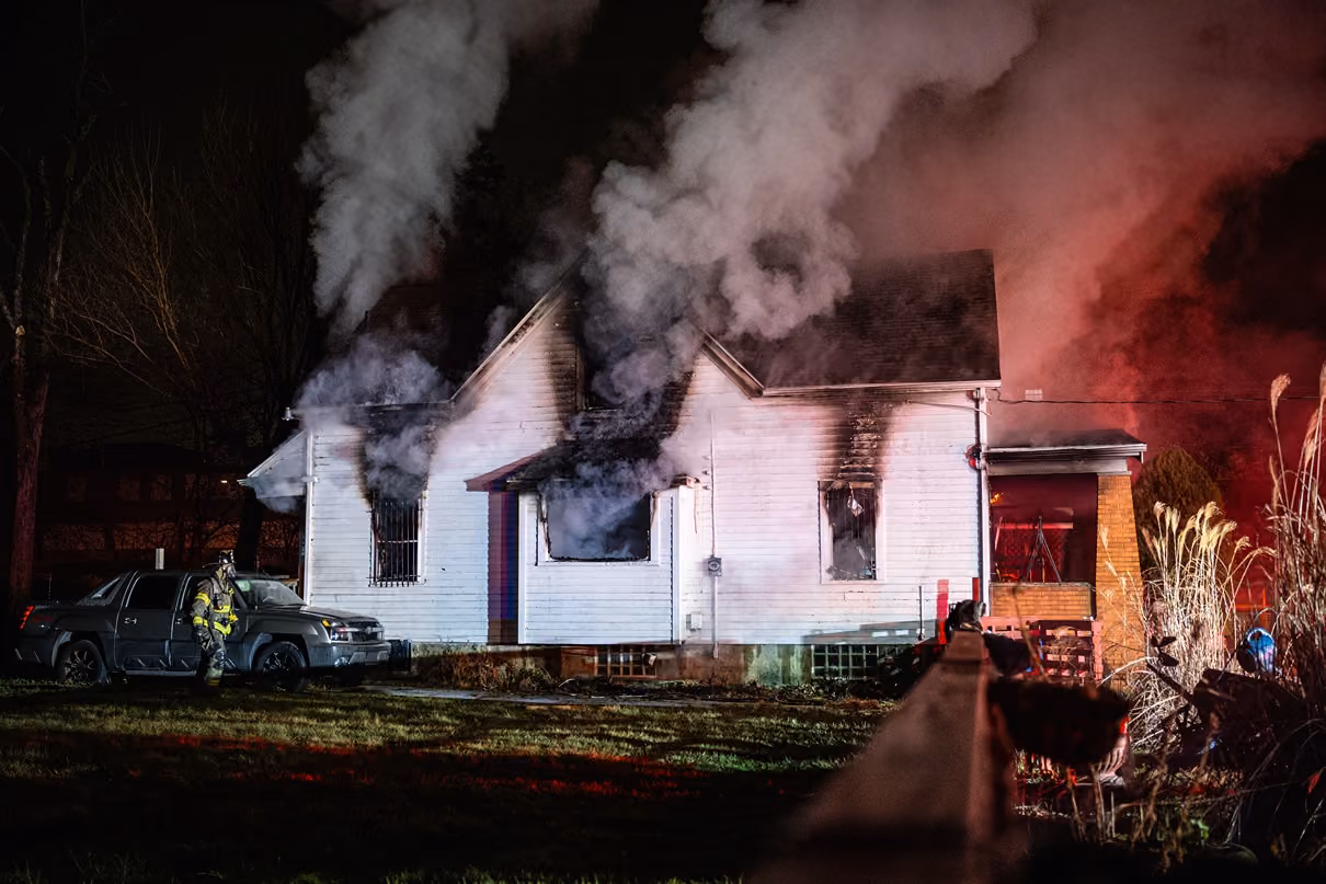 White house emitting thick smoke from windows with firefighter standing near a gray truck at night.