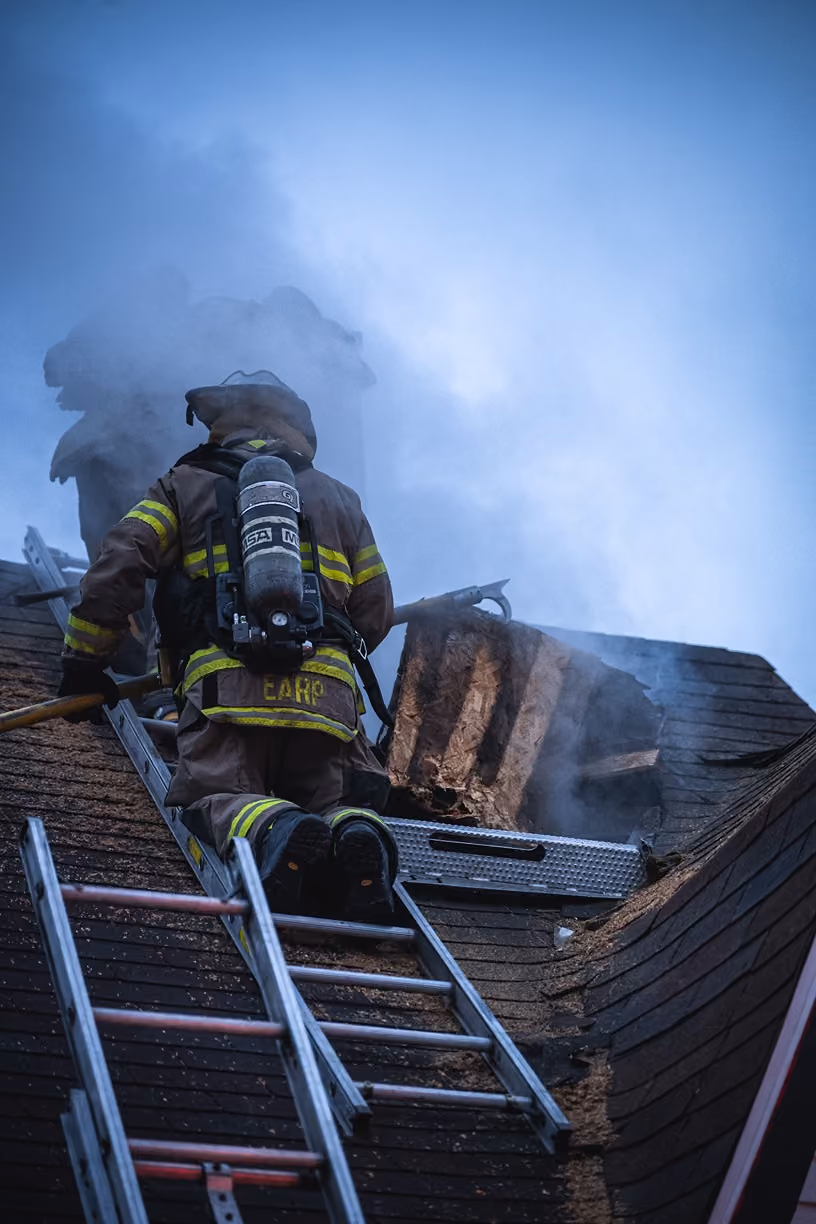 Firefighter in gear climbs roof ladder with smoke rising near damaged chimney at dusk.