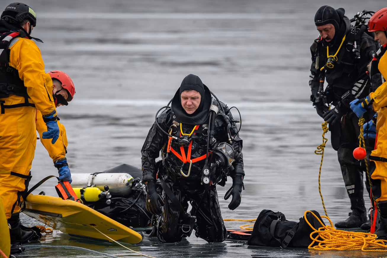 Divers in dry suits and helmets with safety team assisting near water and rescue gear.