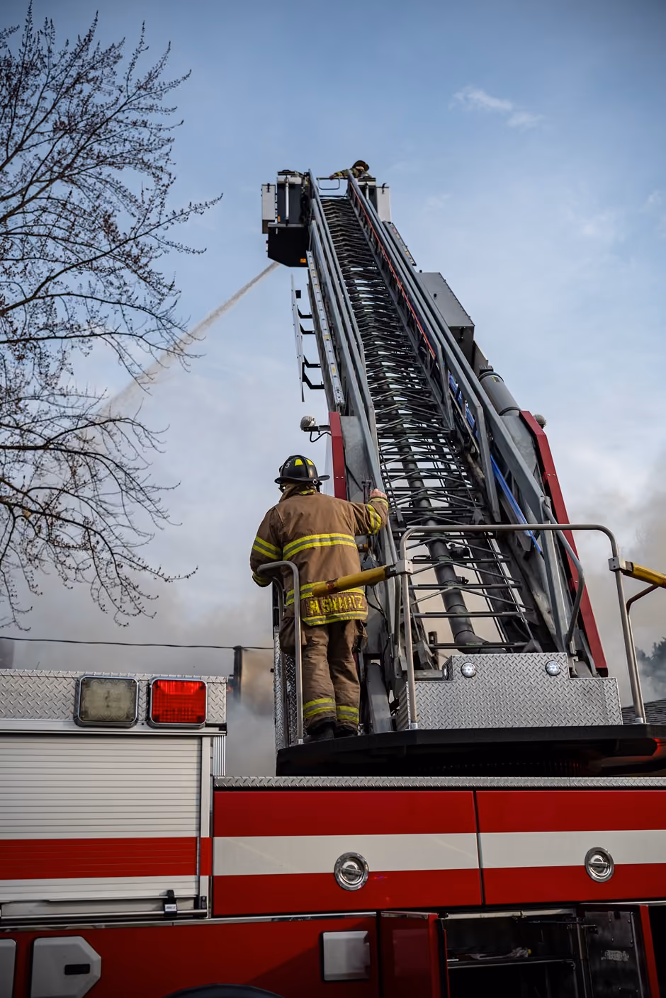 Firefighter in gear on a fire truck's extended ladder spraying water toward a fire.