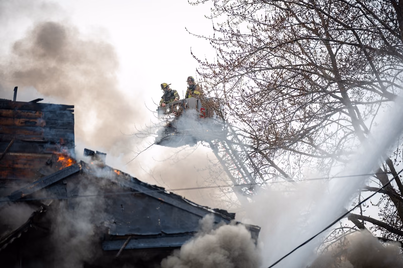 Firefighters on a raised platform spraying water on a burning house with smoke and flames visible.