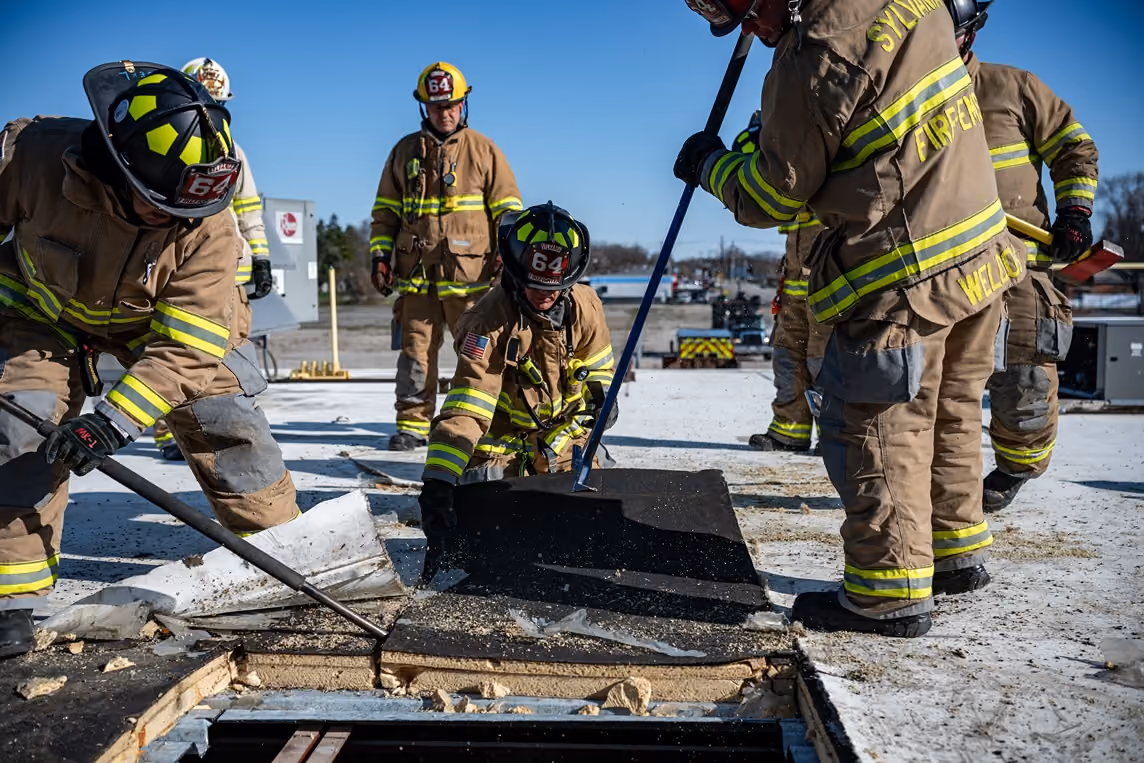 Firefighters in gear removing roofing material from a rooftop ventilation opening under clear sky.