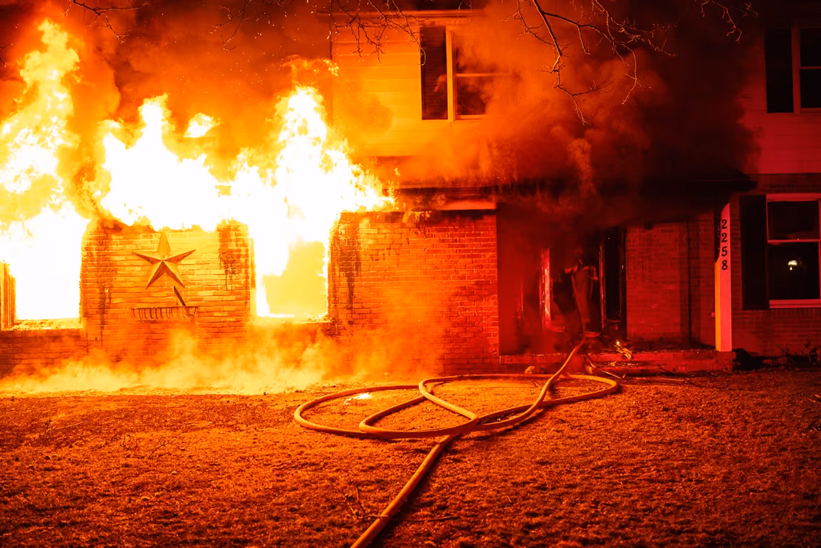 House engulfed in flames at night with fire hose laid on the ground in front.