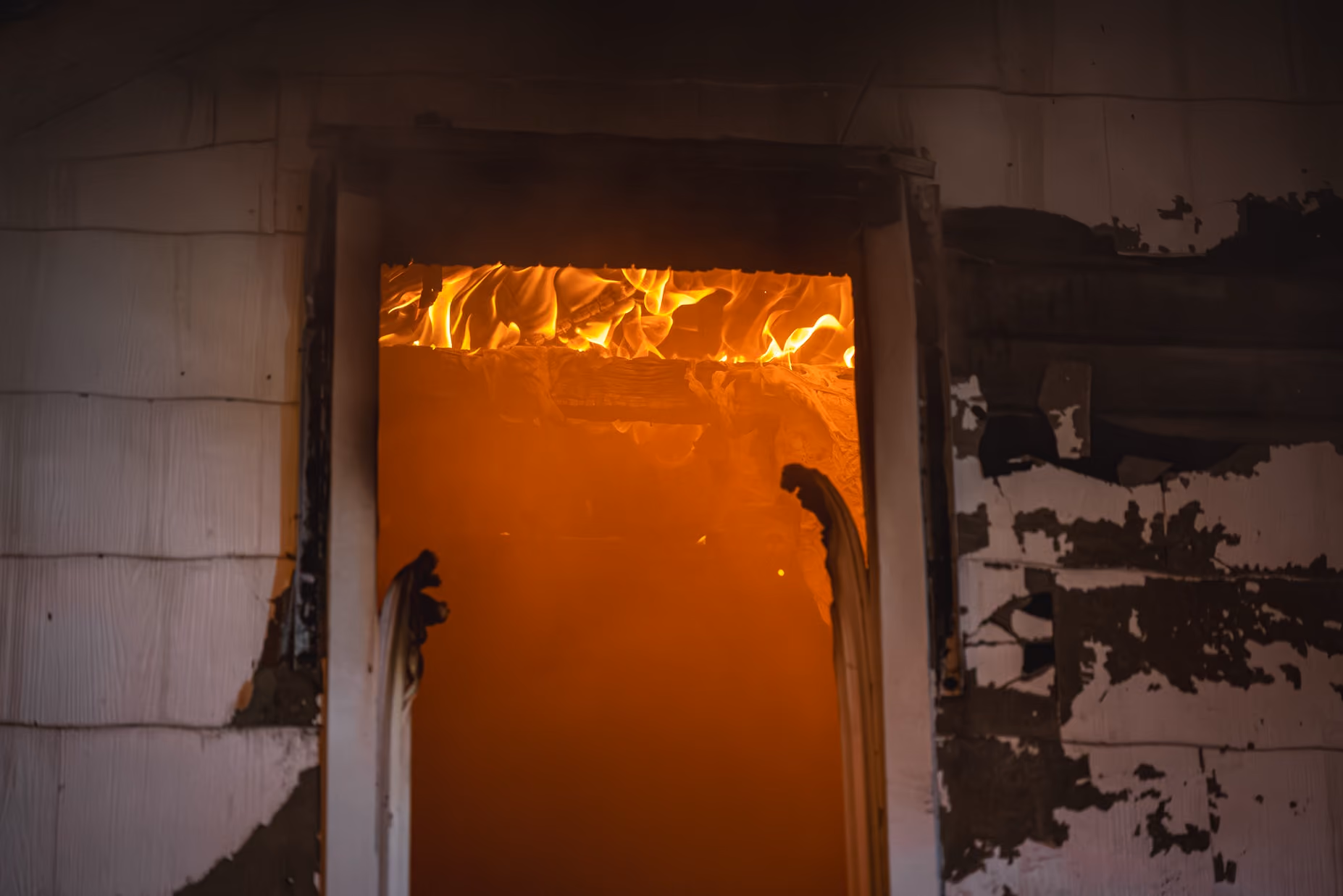 Flames and smoke engulf the interior of a building with charred walls and a burnt door frame.