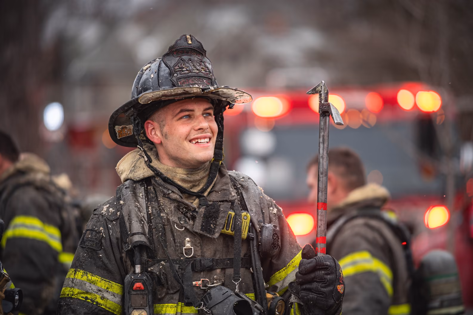 Smiling firefighter in dirty gear holding a tool with fire truck lights blurred in background.