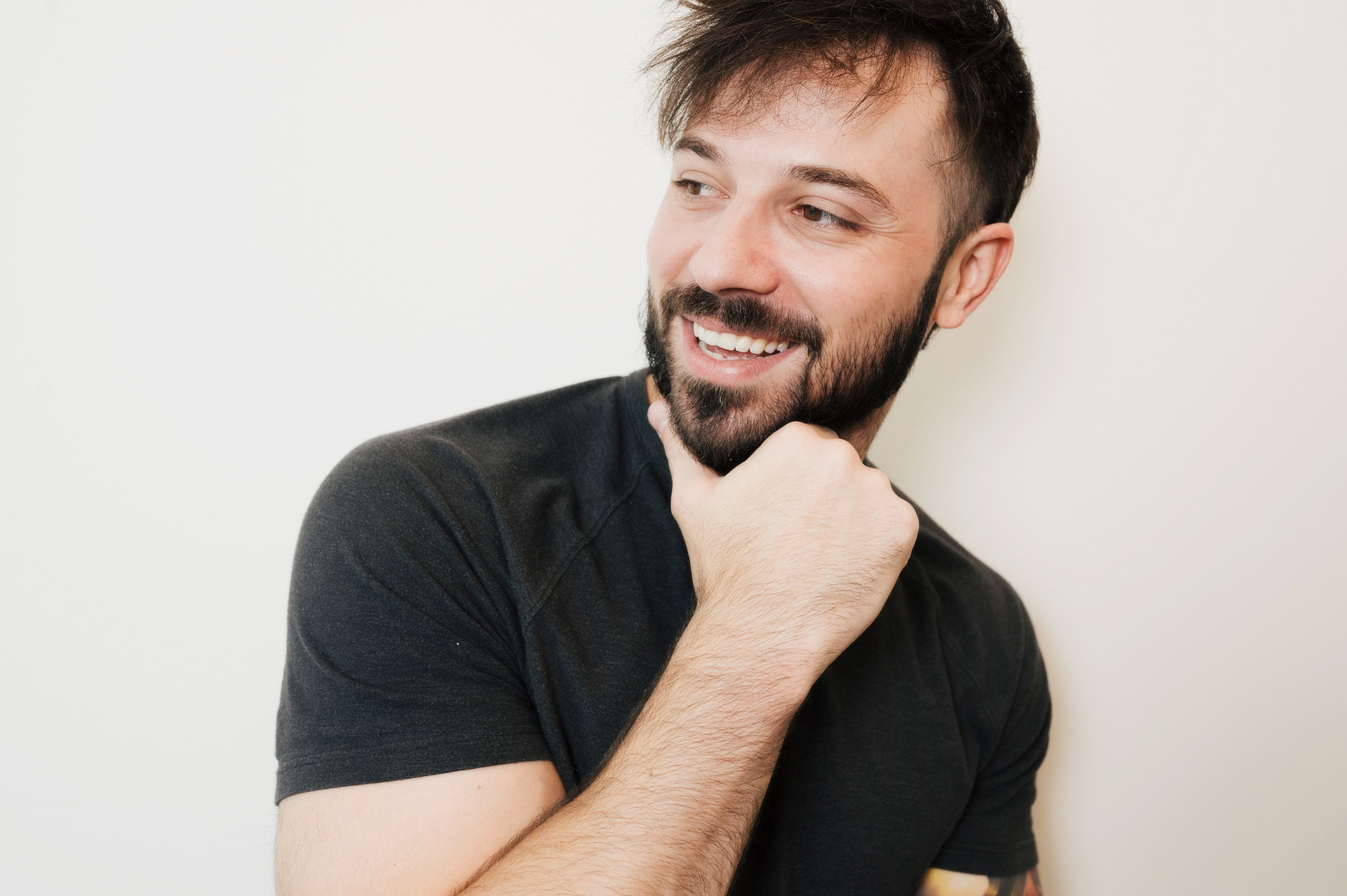 Smiling man with beard wearing a black t-shirt resting his chin on his hand against a plain white background.