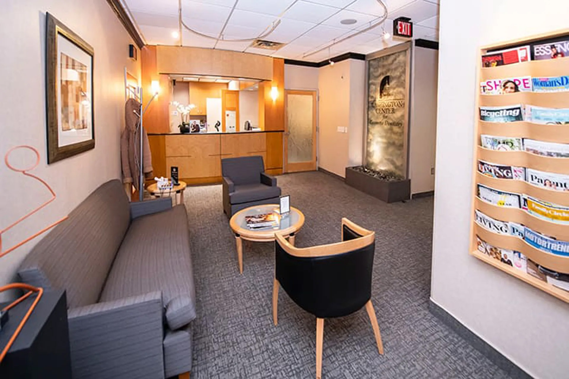 Reception area with gray seating, round table, magazine rack, and wooden front desk.
