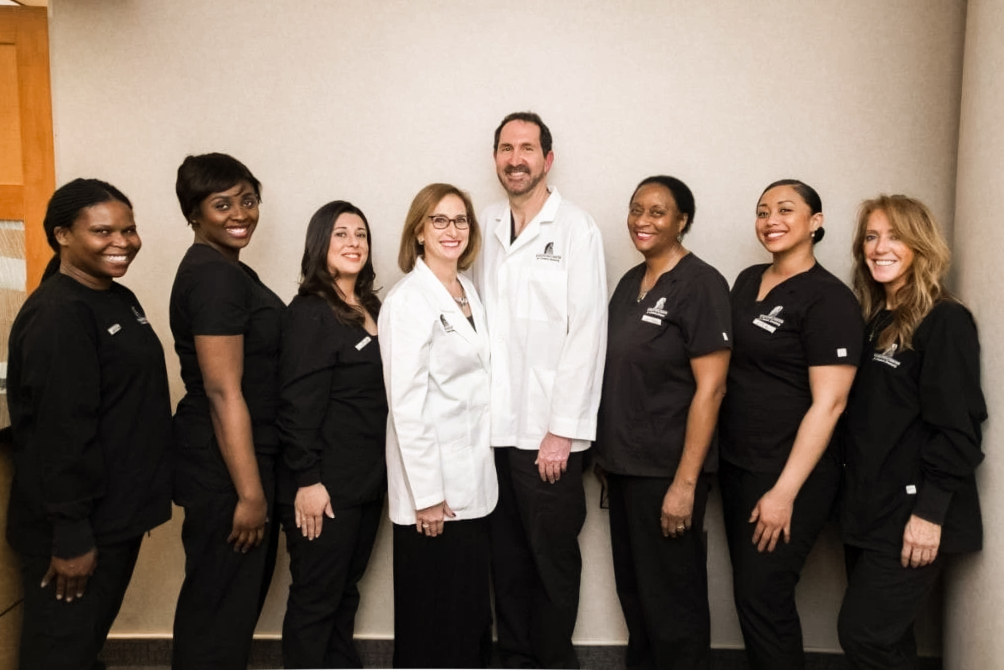 Group of eight diverse medical staff, two in white coats and six in black scrubs, posing and smiling indoors.