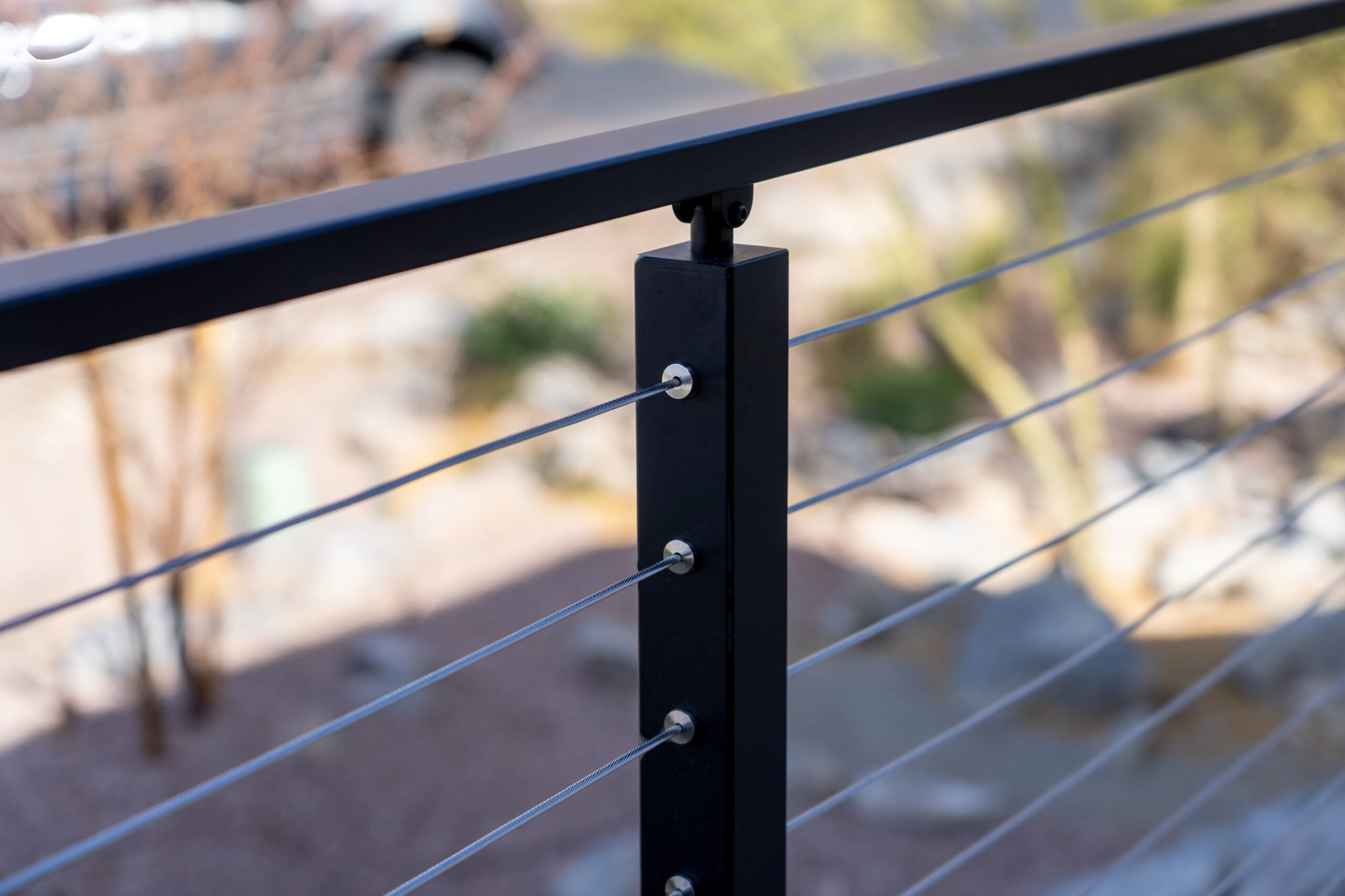 Close up of the handrail, post, and cable of an exterior Signature Cable Railing system with a sunny, blurred background