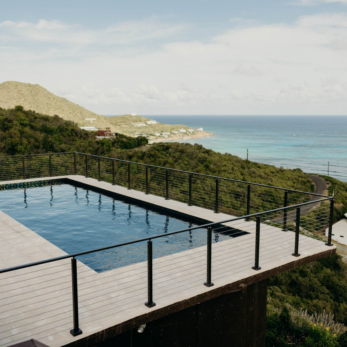 A pool terrace edged in black rod railing sits above a green headland