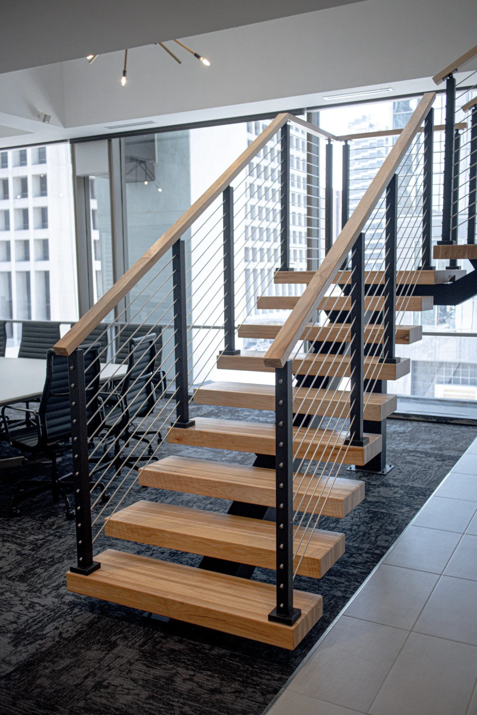 Office stair with light treads and cable railing beside a glass conference room and city towers
