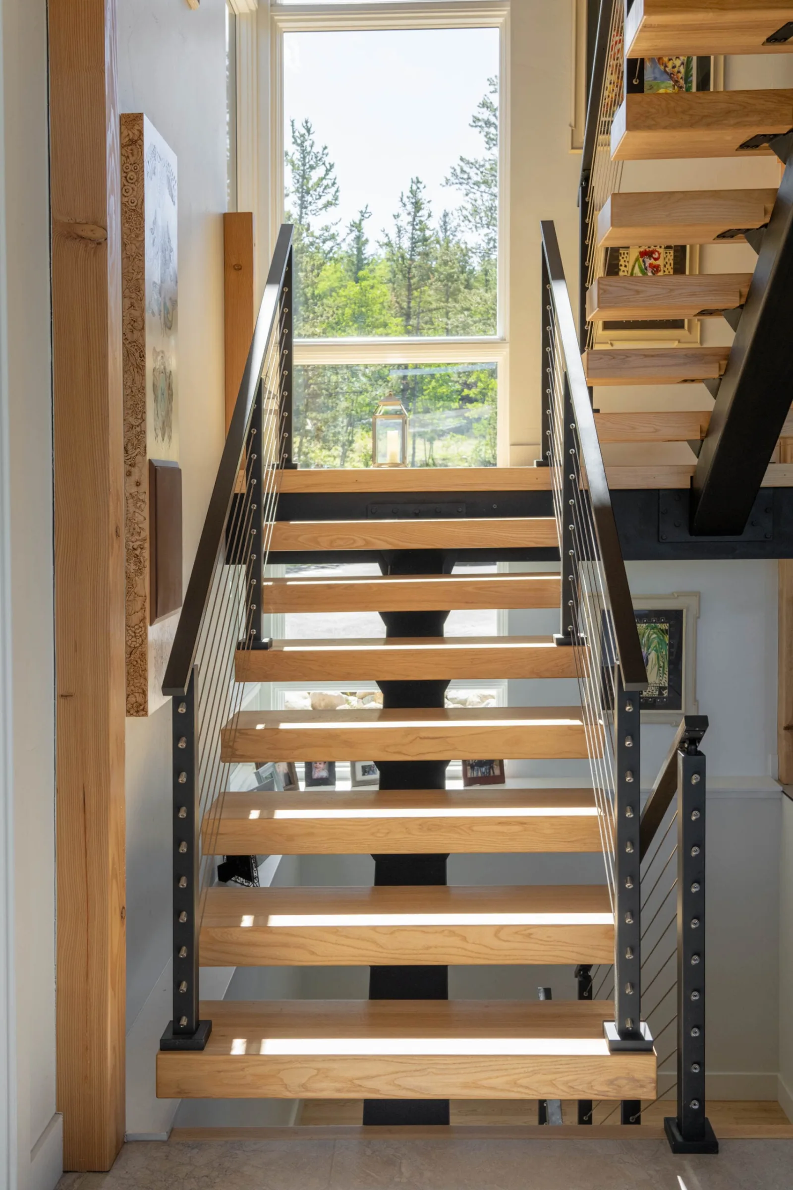 Straight stair of wood treads with black cable railing rising toward a window of evergreens