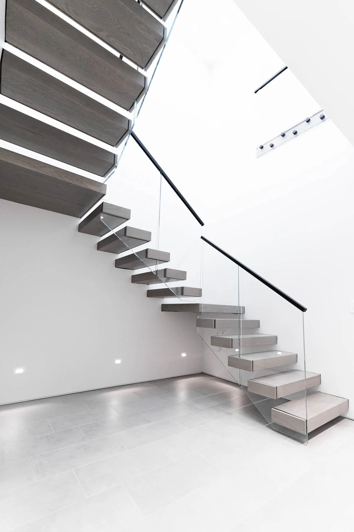 Cantilevered gray treads springing from a white wall with glass railing and slim black handrail