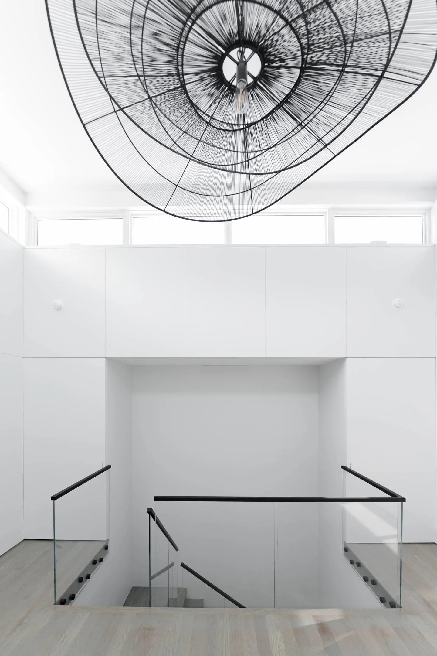Double-height white room with a sculptural pendant above a glass-railed stair opening