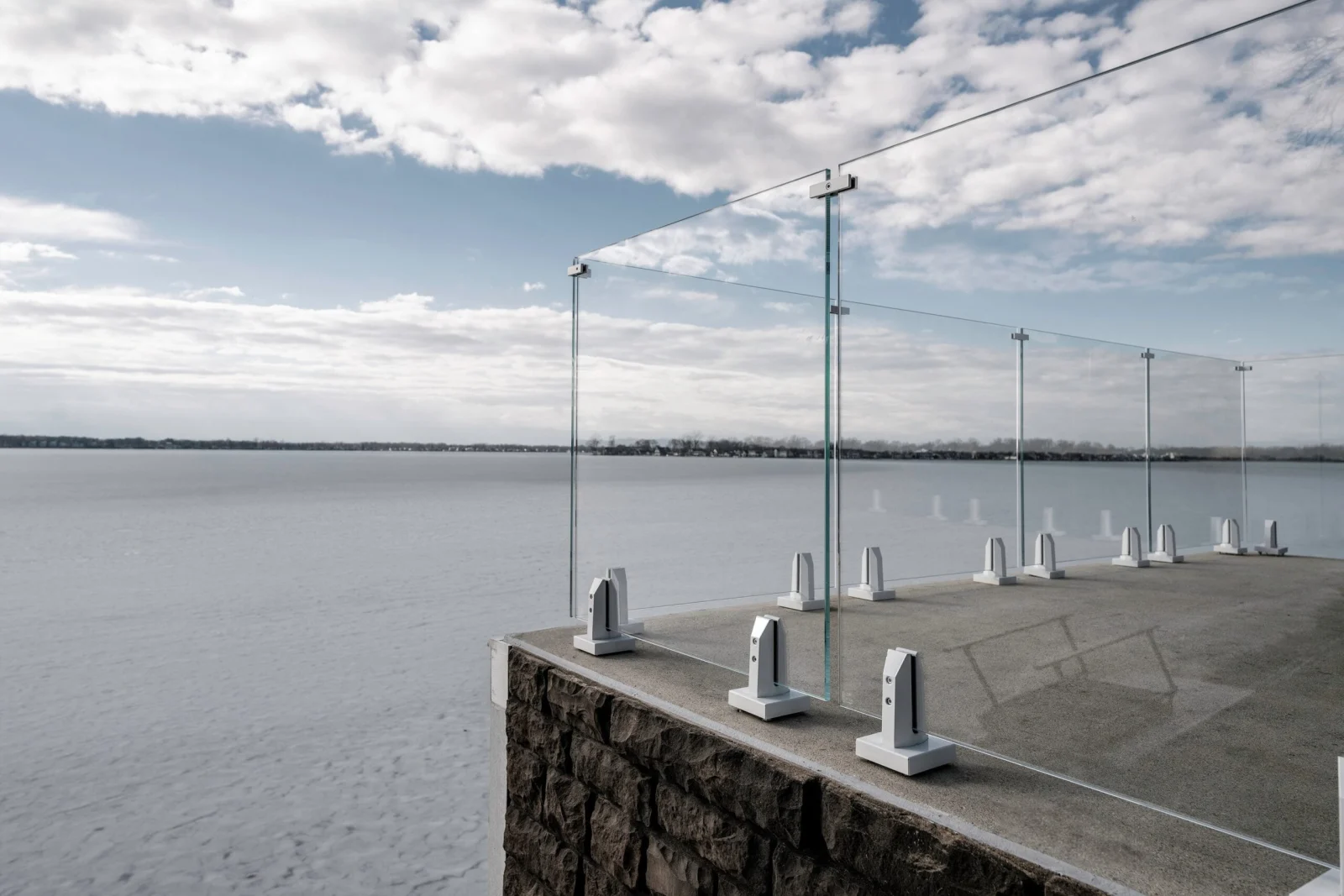 Frameless glass panels in metal base clamps along a concrete terrace above a frozen lake