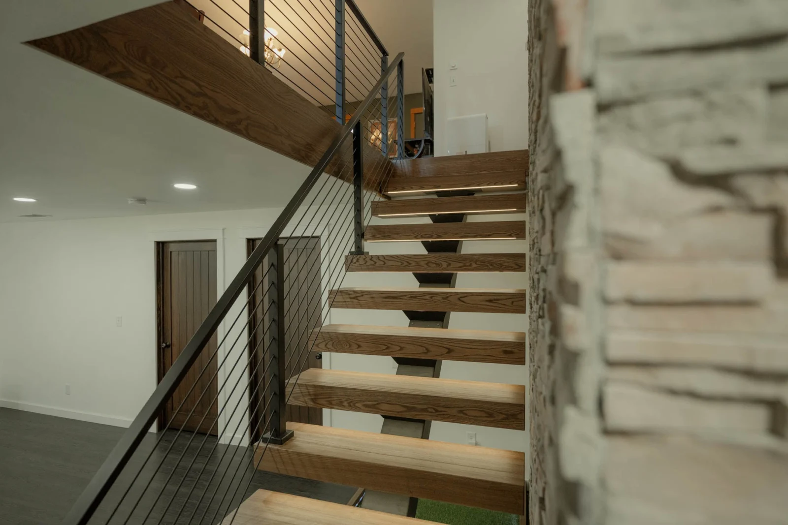 Open-riser wood treads on a dark center stringer with black rod railing beside stacked stone