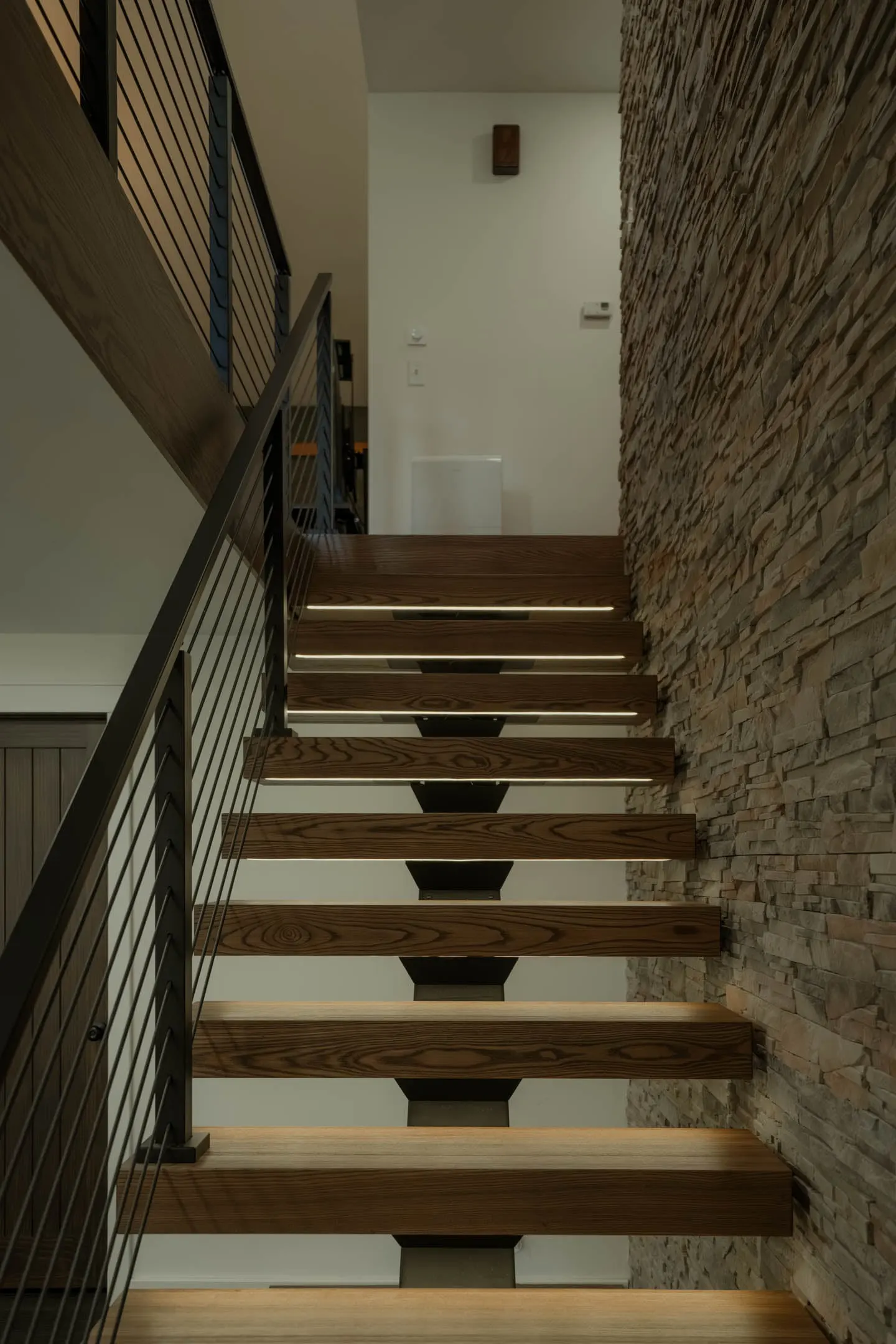 View up the flight with lit wood treads and rod railing beside stacked stone