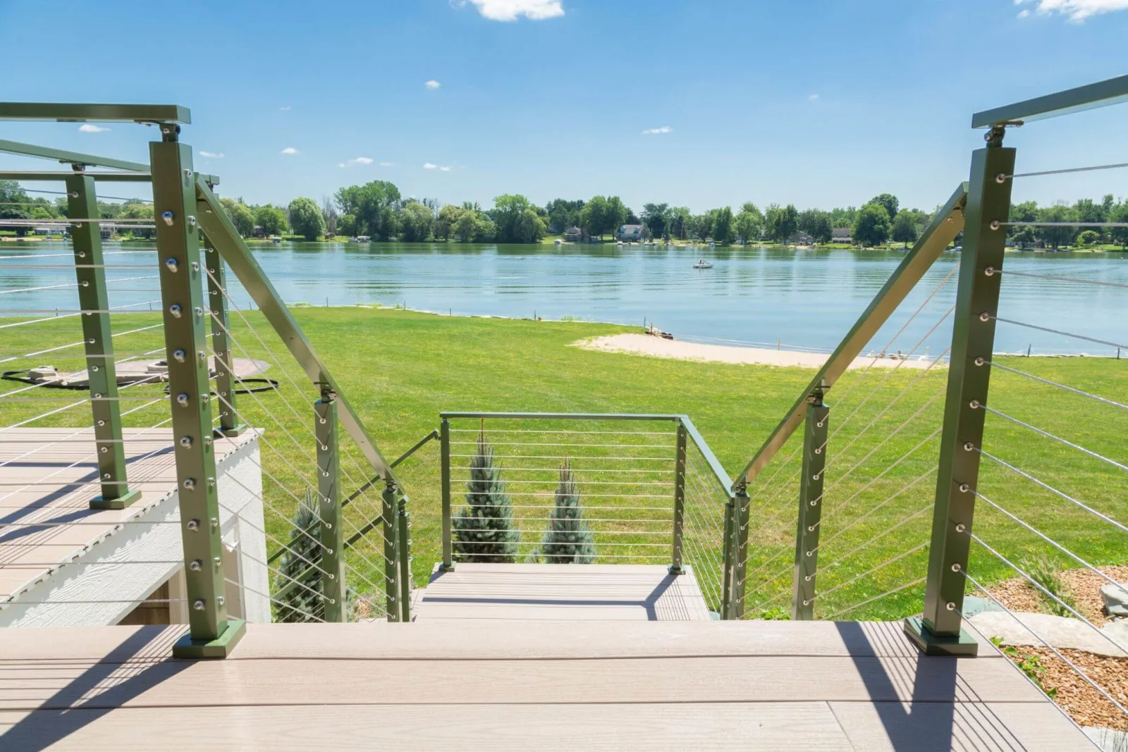 Deck steps descending toward a lawn and lake between green powder-coated railing