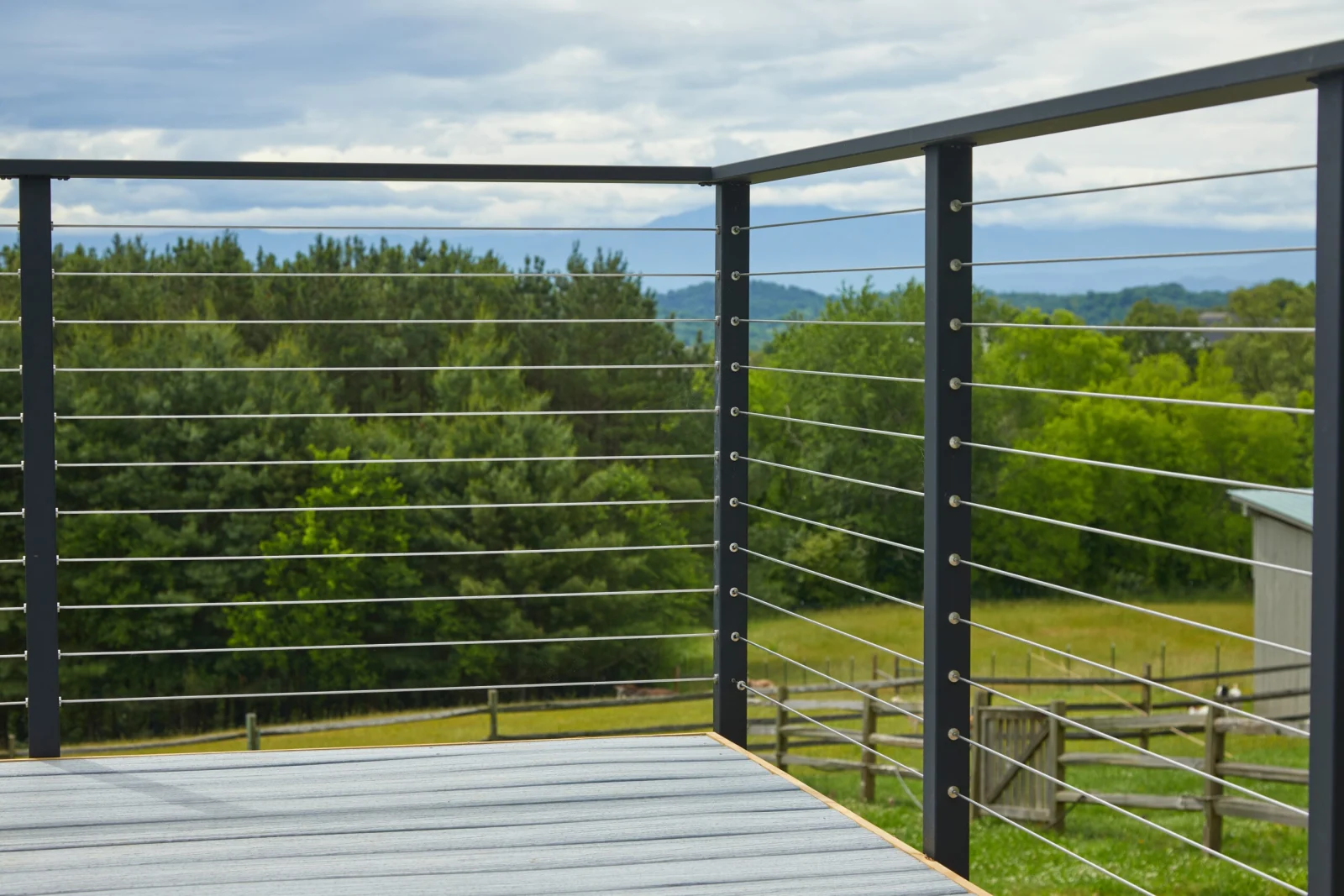 Corner of a gray deck framed by black cable railing facing trees and open pasture
