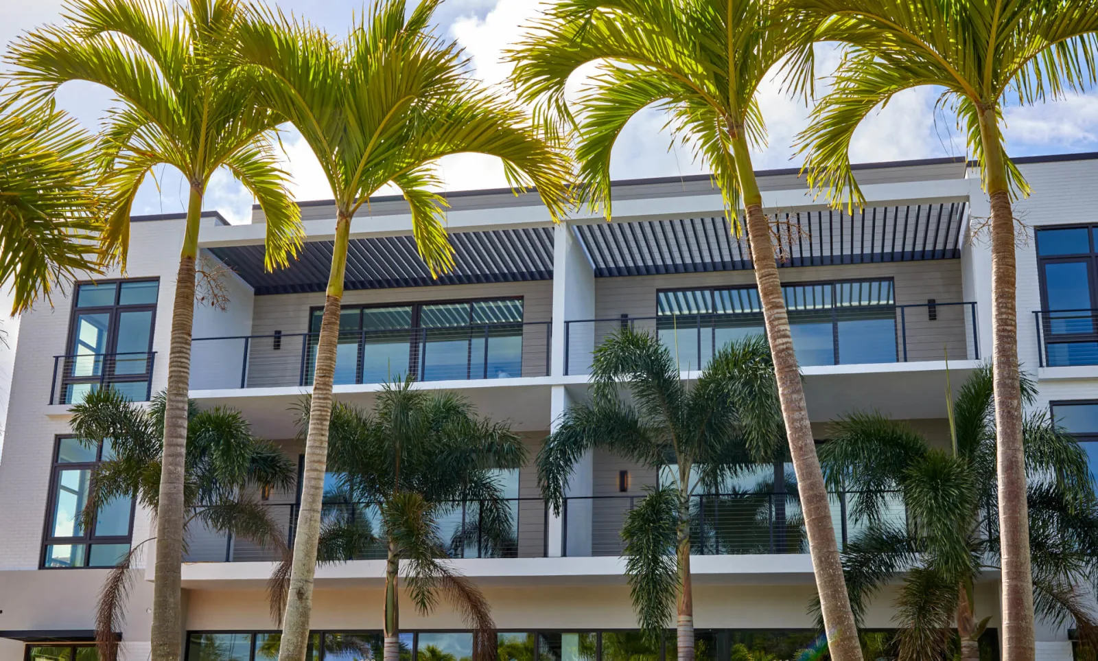 Three-story condo building behind palms with black cable railing on each balcony