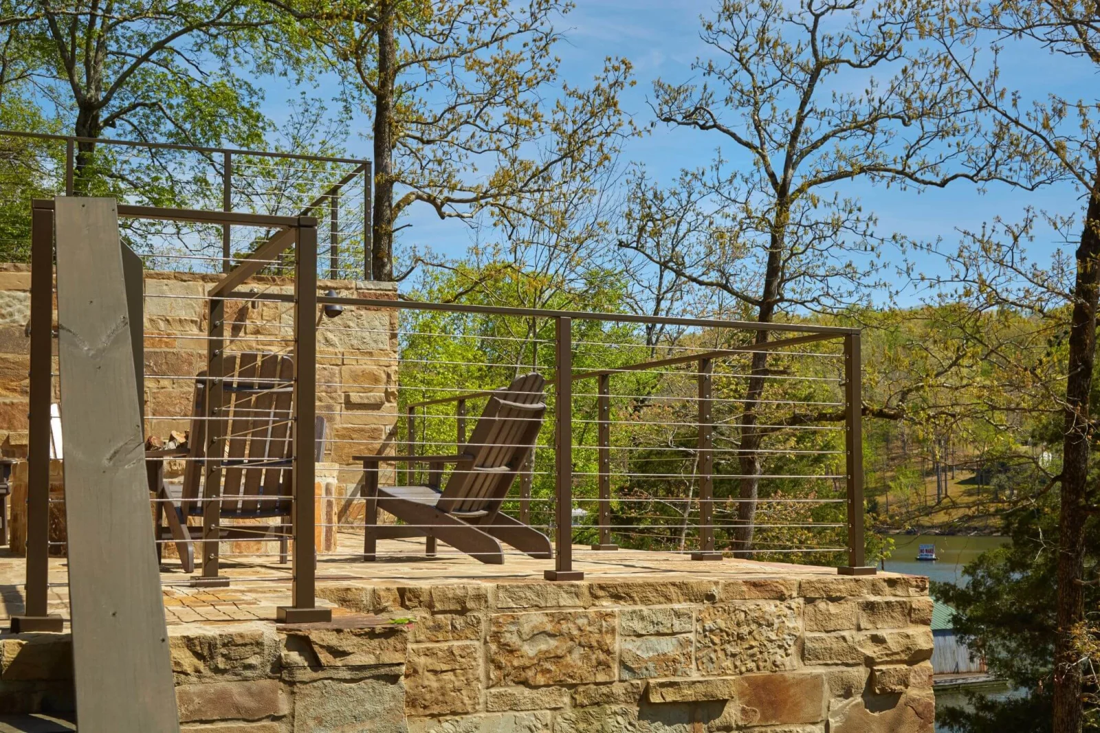 Bronze cable railing along a stone patio with wood chairs above a lake