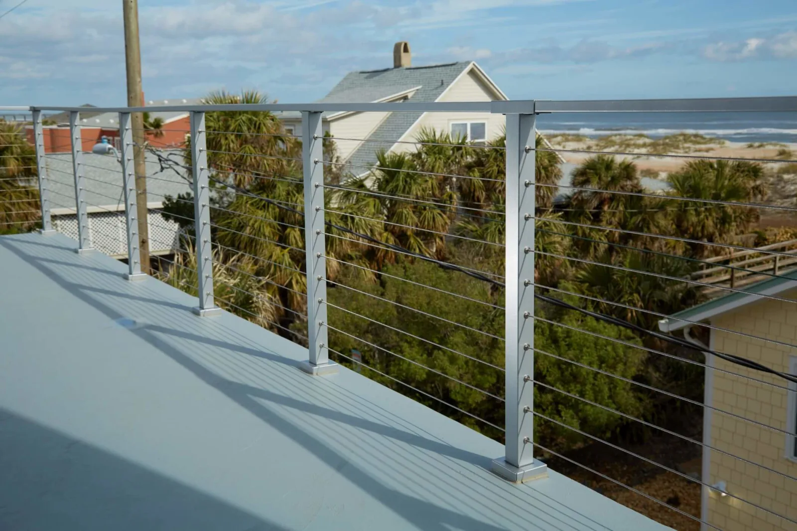Flat-top silver posts and cable ringing a rooftop deck above palms with ocean beyond