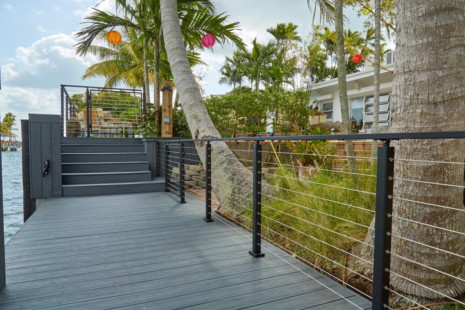 Waterside deck with black cable railing curving past a palm trunk toward steps