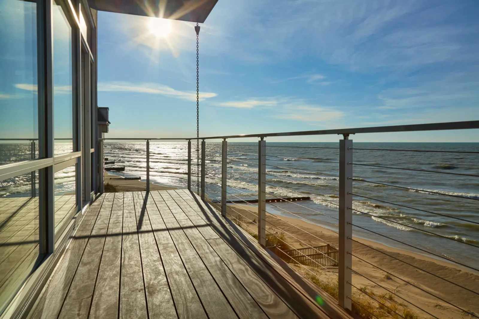 Sun flare over a weathered plank balcony with silver cable railing facing a beach