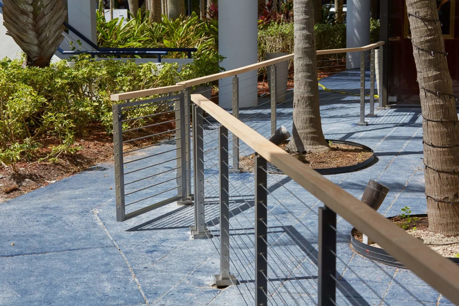 Wood-capped cable railing lining a sloped walkway between palm trunks on blue pavers