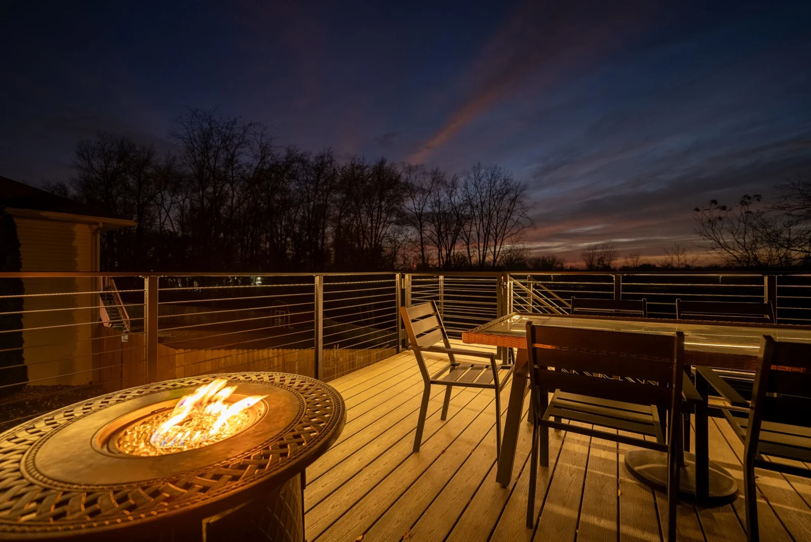 Dusk on the deck with a fire table, dining chairs and rod railing silhouette