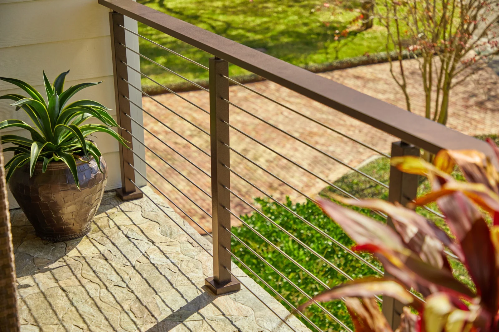 Copper-toned railing with horizontal rods and wide top rail edging a stone porch and garden